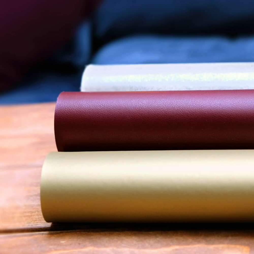 Visual of three rolls of material on a wooden table, 1 is gold, 1 is magenta, 1 is silver.