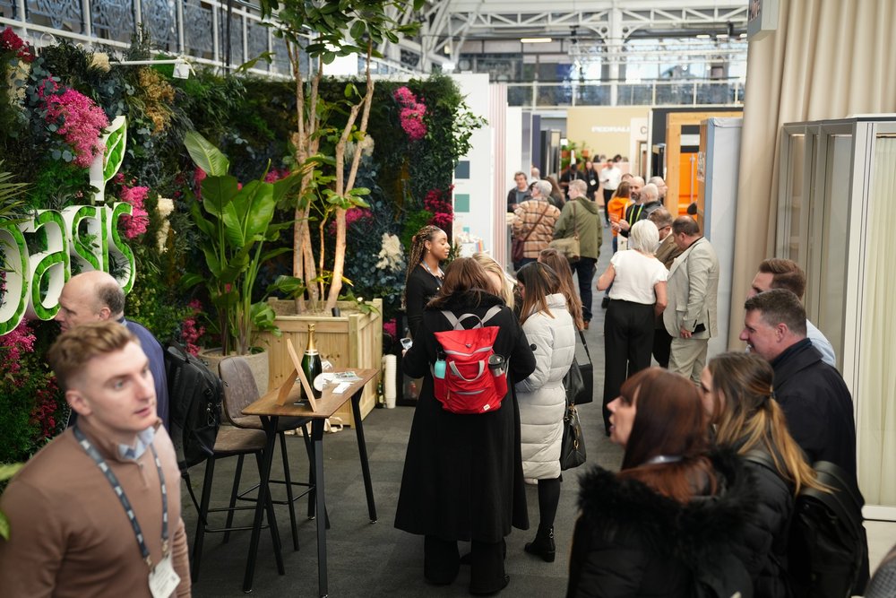 People walk the floor of the exhibitor stands in the workspace design show. One wall is lined with green leaves and plants to the left.