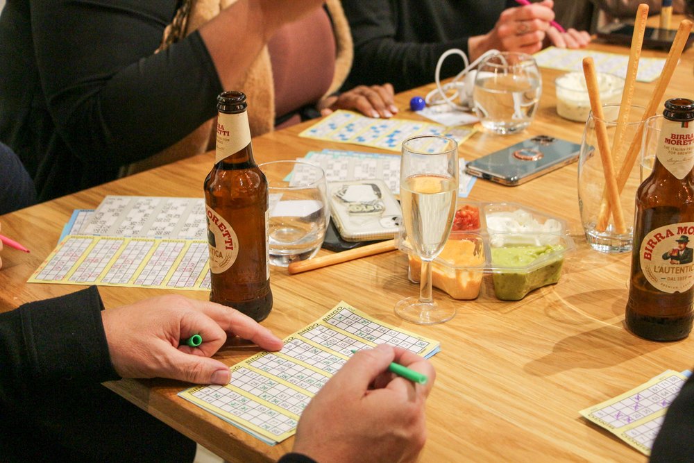 A beer bottle and glasses sit atop a table, where people are playing a game of bingo. A right hand holds a pen over a bingo card.