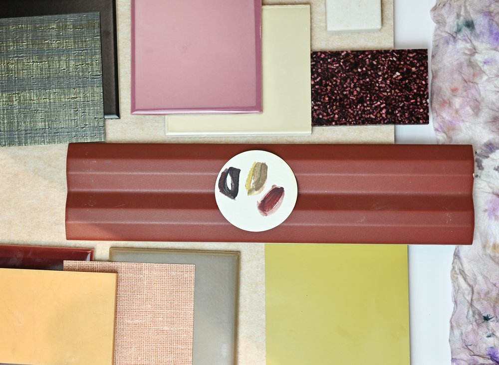 An image of tiles, materials, fabrics and a paint palette in various colours are laid out on a flat surface. The materials are layered over each other to create a moodboard or flatlay.