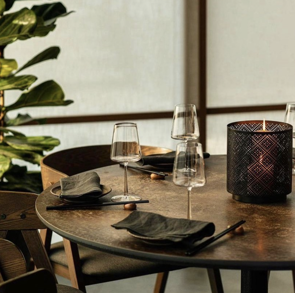 A circular wooden table sits in the centre, surrounded by wooden chairs. The table is set with napkins, chopsticks and wine glasses, and a large metal candle holder sits atop the centre of the table.