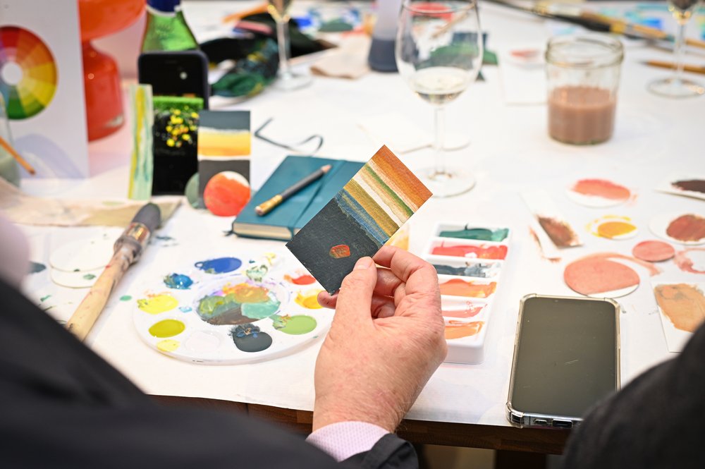 An image of a hand holding a piece of paper with different colour tones painted on. The table has paint palettes and paint brushes with different colour swatches.