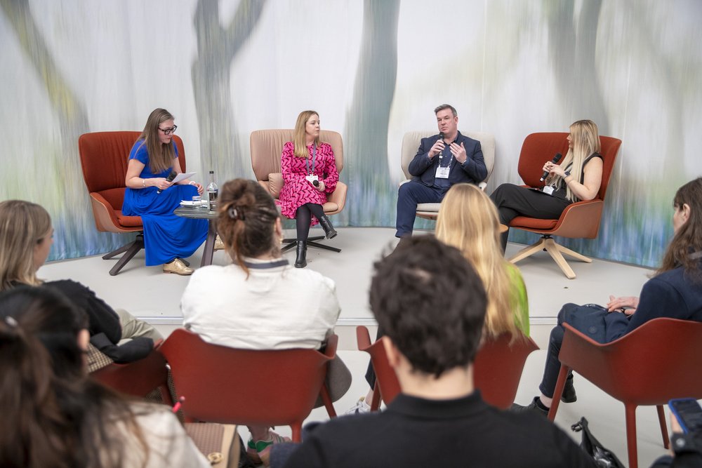 Four people sit on armchairs on stage, in the foreground is the backs of the audience heads.