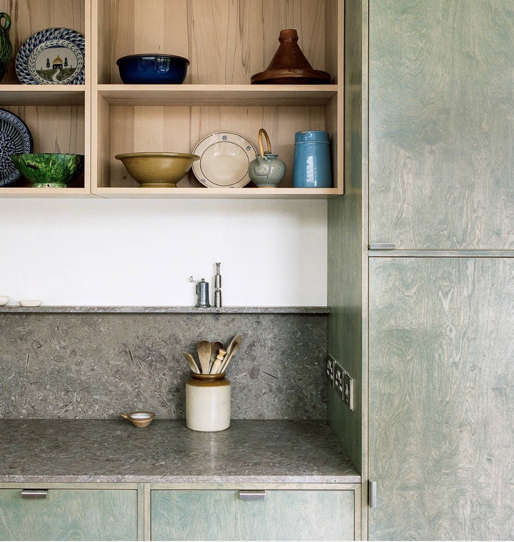 Image of green paint stained kitchen cupboards - with plywood shelving and kitchen utensils on a granite worktop.